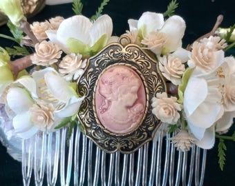 May include: A silver hair comb with a cameo featuring a woman's profile in pink. The comb is decorated with white and cream artificial flowers.