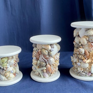May include: Three white round pedestals of varying sizes, each decorated with a different arrangement of seashells. The pedestals are arranged from smallest to largest, with the largest pedestal in the back.