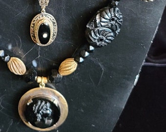 May include: A black beaded necklace with three pendants. The pendants are made of black and gold metal and feature cameo-like designs. One pendant has a black cameo of a woman's profile, another has a black cameo of a floral design, and the third has a brown cameo of a floral design.