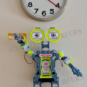 May include: A robot made of gray and yellow plastic with large eyes and a Meccano logo on its chest. The robot is holding a 50 Australian dollar bill in its hand. A clock with a white face and black numbers is on the wall behind the robot.