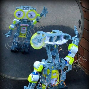 May include: Two robots made of grey and yellow plastic with wires and gears visible. The robot on the left is reflected in a mirror. The robot on the right has a large round wheel on its back and is standing on a black surface.