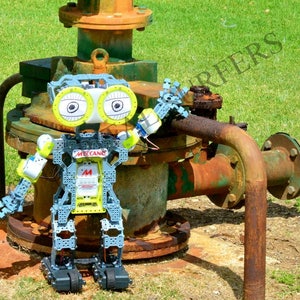 May include: A robot made of gray plastic with yellow accents and large, round eyes. The robot is standing on a rusty metal pipe and valve. The robot is labeled "Meccano" on its chest.