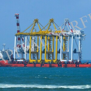 Może przedstawiać: Czerwono-biały statek towarowy z dużym żółto-białym dźwigiem na pokładzie. Statek płynie po błękitnym oceanie z białymi falami.