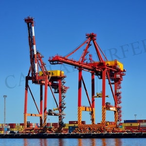 Może przedstawiać: Czerwone i żółte dźwigi przemysłowe z żółto-czarnymi pasami ostrzegawczymi w porcie. Dźwigi służą do załadunku i rozładunku statków towarowych.