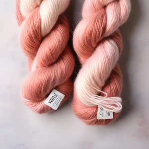 May include: Two twisted skeins of yarn featuring a gradient color pattern that transitions from a deep dusty rose pink to a soft creamy white. Each skein includes a small white paper label with black printed text attached to the bottom.