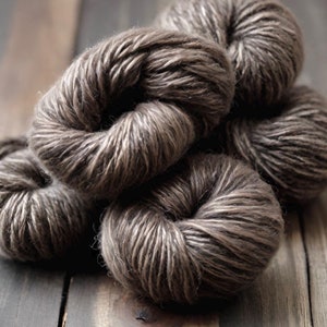 May include: Close-up of several skeins of brown yarn. The yarn is tightly wound into round balls, with a mix of light and dark brown fibers. The yarn is arranged on a weathered wooden surface, creating a rustic aesthetic.