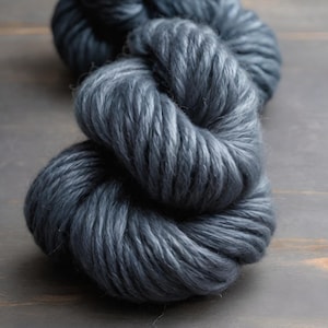 May include: Close-up of two skeins of thick, dark gray yarn. The yarn is tightly wound, with visible texture and a slight sheen. The background is a dark, neutral color, highlighting the yarn's texture. Ideal for knitting or crocheting projects.
