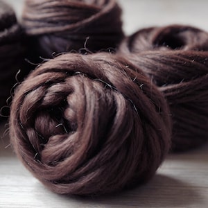 May include: Close-up of several dark brown yarn balls, tightly wound and textured. The yarn appears soft and fluffy, with a slight sheen. The balls are arranged on a light-colored wooden surface, suggesting a craft or textile theme.