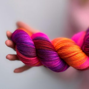 Może przedstawiać: Motka włóczki w odcieniach fioletu, różu i pomarańczy. Włóczka jest skręcona i spleciona, tworząc kolorowy i fakturowany wygląd.