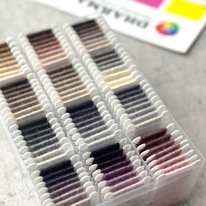 May include: A clear plastic organizer filled with spools of thread in various shades of gray, brown, and purple. A color chart with the word "DHARMA" is visible in the background, with a rainbow-colored logo.