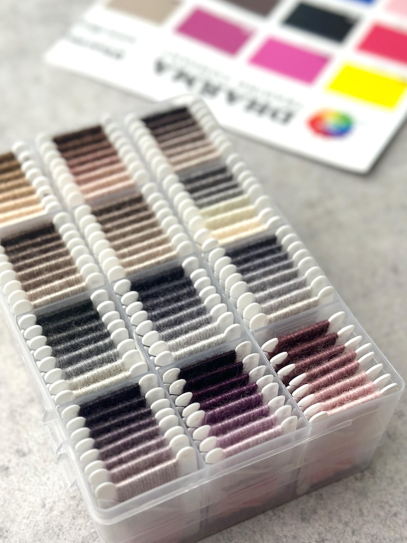 May include: A clear plastic organizer filled with spools of thread in various shades of gray, brown, and purple. A color chart with the word "DHARMA" is visible in the background, with a rainbow-colored logo.
