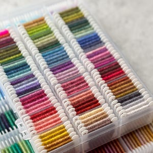 May include: A clear plastic organizer filled with spools of colorful embroidery floss. The floss is arranged in rows, showcasing a spectrum of colors from vibrant pinks and yellows to deep blues and greens. Each spool is labeled with a number.