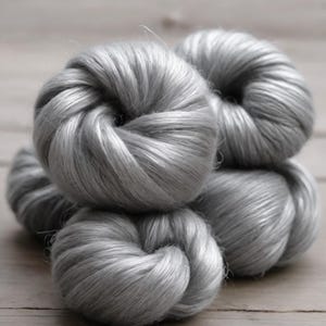 May include: Close-up of several skeins of silver-gray yarn, tightly wound into rounded shapes. The yarn has a soft, silky texture, and the lighting highlights the subtle variations in color and texture. The yarn is likely used for knitting or crocheting.