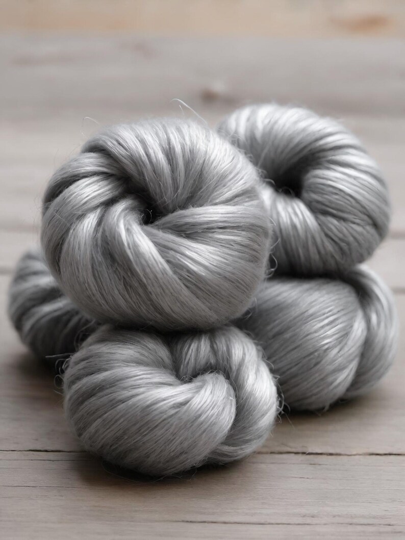 May include: Close-up of several skeins of silver-gray yarn, tightly wound into rounded shapes. The yarn has a soft, silky texture, and the lighting highlights the subtle variations in color and texture. The yarn is likely used for knitting or crocheting.