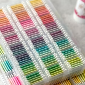 May include: A close-up of several clear plastic containers filled with colorful embroidery floss. The floss is arranged in neat rows, showcasing a spectrum of colors from yellow to purple and green. The containers are stacked, suggesting a large collection.