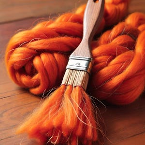May include: A wooden-handled paintbrush with a silver ferrule rests on a pile of vibrant orange wool roving. The brush's bristles are saturated with the same bright orange color. The scene is set on a warm, wooden surface, creating a contrast of textures.