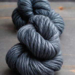 May include: A close-up shot of a skein of thick, dark gray yarn. The yarn is tightly wound and has a soft, textured appearance. The color is a deep, muted gray, with subtle variations in tone. The yarn is likely used for knitting or crocheting projects.
