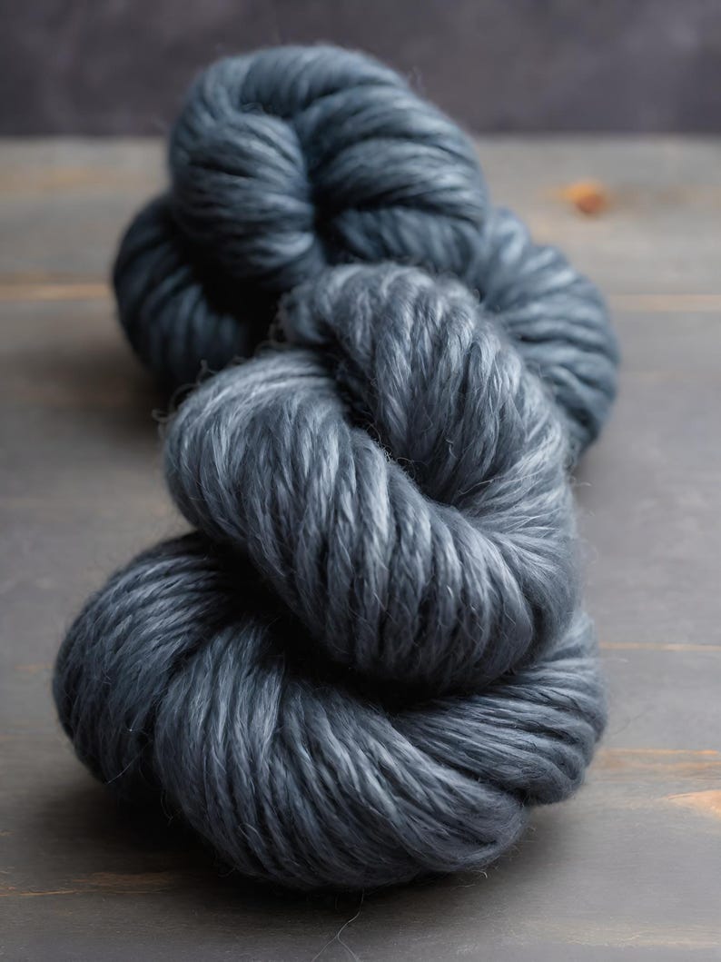 May include: A close-up shot of a skein of thick, dark gray yarn. The yarn is tightly wound and has a soft, textured appearance. The color is a deep, muted gray, with subtle variations in tone. The yarn is likely used for knitting or crocheting projects.