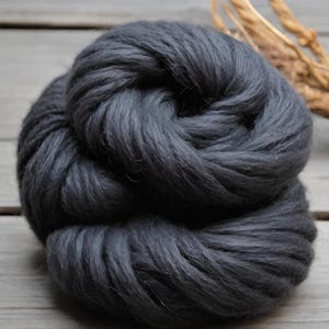 May include: A large, dark gray ball of thick yarn, tightly wound. The yarn appears soft and fluffy, with a slightly textured surface. The background is a light gray wooden surface. A bundle of dried, light brown plant stems is in the background.