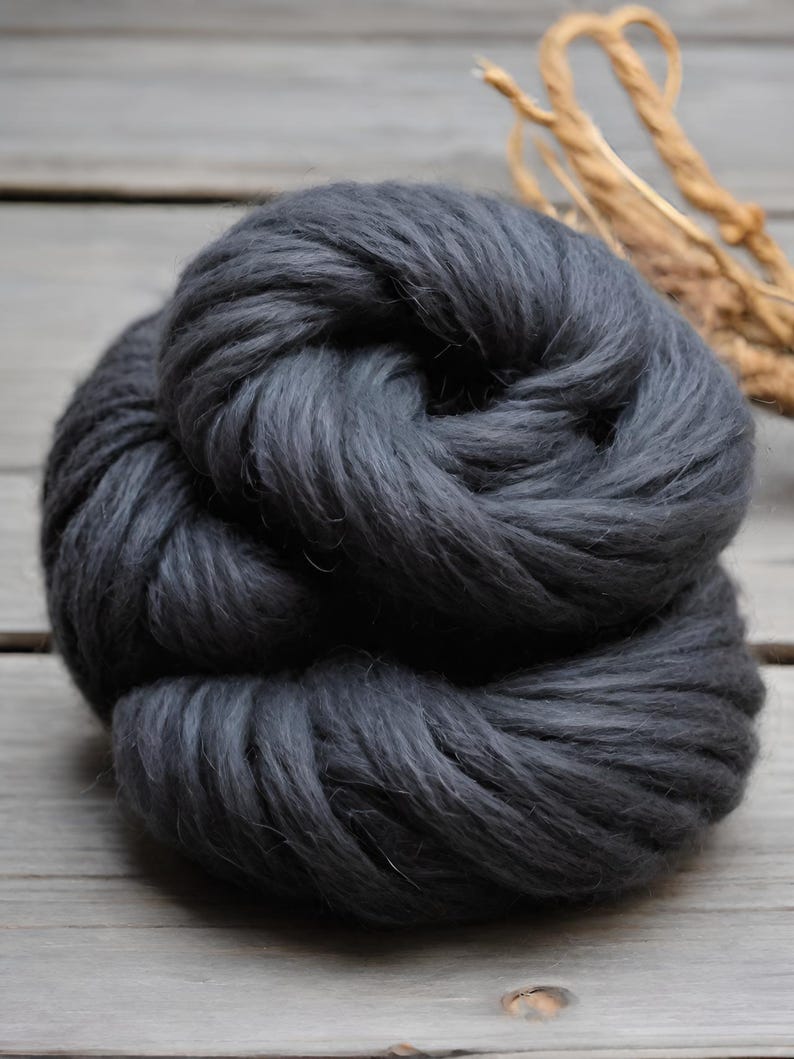 May include: A large, dark gray ball of thick yarn, tightly wound. The yarn appears soft and fluffy, with a slightly textured surface. The background is a light gray wooden surface. A bundle of dried, light brown plant stems is in the background.