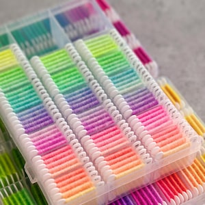 Könnte beinhalten: Eine Sammlung farbenfroher Stickgarne, die in transparenten Kunststoff-Aufbewahrungsboxen organisiert sind. Die Garne sind nach Farben sortiert, von leuchtenden Grün- und Gelbtönen bis hin zu Rosa, Lila und Blau. Jede Box ist in Abschnitte unterteilt, in denen die Garne ordentlich aufgewickelt sind.
