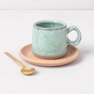 May include: A light green ceramic coffee cup with a speckled glaze sits on a beige saucer with a speckled glaze. A gold spoon is placed to the left of the saucer.