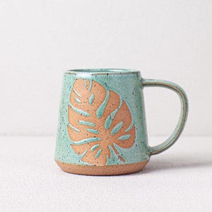 May include: A teal green ceramic mug with a brown leaf design. The mug has a speckled glaze and a rounded shape.