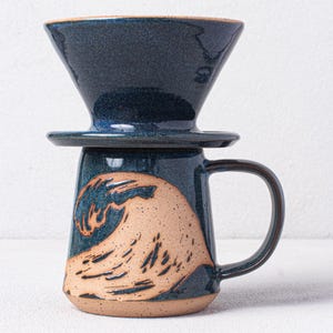 May include: A blue ceramic coffee mug with a wave design. The mug has a matching blue ceramic pour-over coffee filter on top.