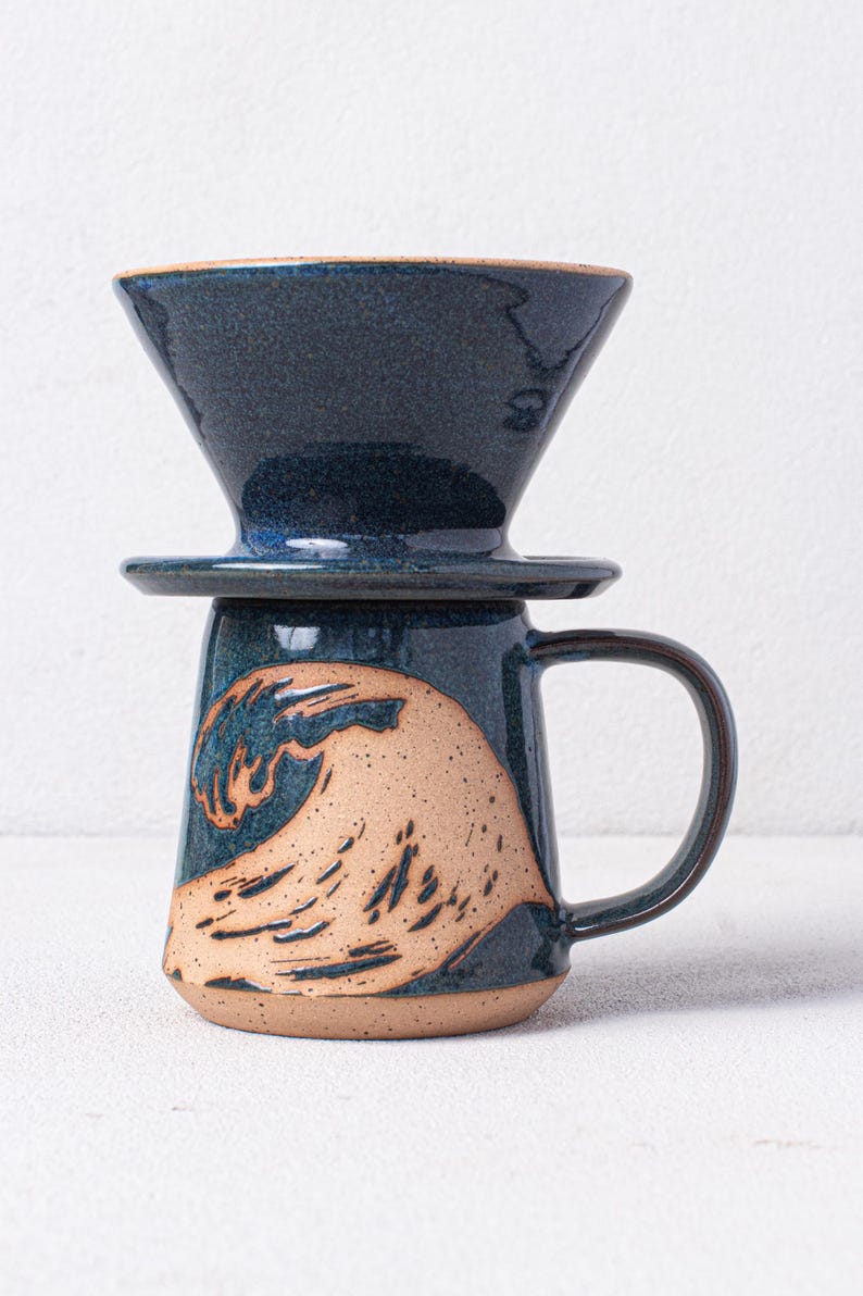 May include: A blue ceramic coffee mug with a wave design. The mug has a matching blue ceramic pour-over coffee filter on top.