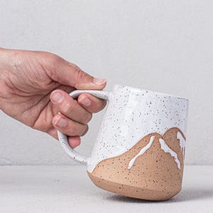 May include: A white ceramic mug with a speckled glaze and a brown mountain design. The mug has a rounded shape and a handle.