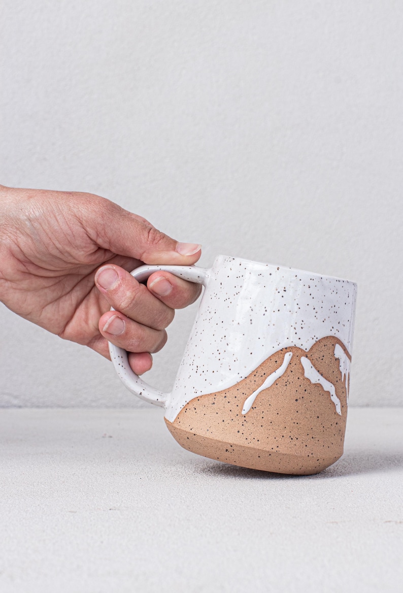 May include: A white ceramic mug with a speckled glaze and a brown mountain design. The mug has a rounded shape and a handle.