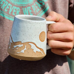 May include: White ceramic mug with a brown mountain and sun design. The mug has a speckled glaze and a rounded handle.