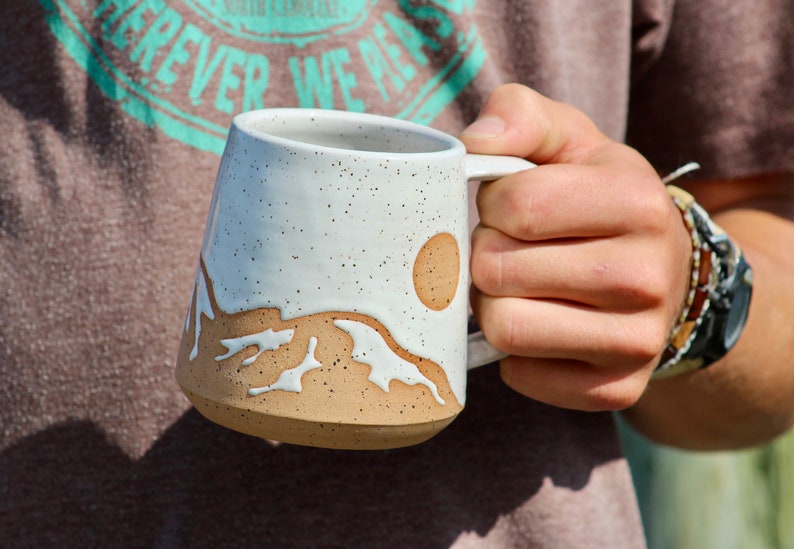 May include: White ceramic mug with a brown mountain and sun design. The mug has a speckled glaze and a rounded handle.
