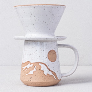 May include: White ceramic coffee pour over with a brown speckled glaze and a brown and white mountain design. The pour over sits on a matching mug with a handle.