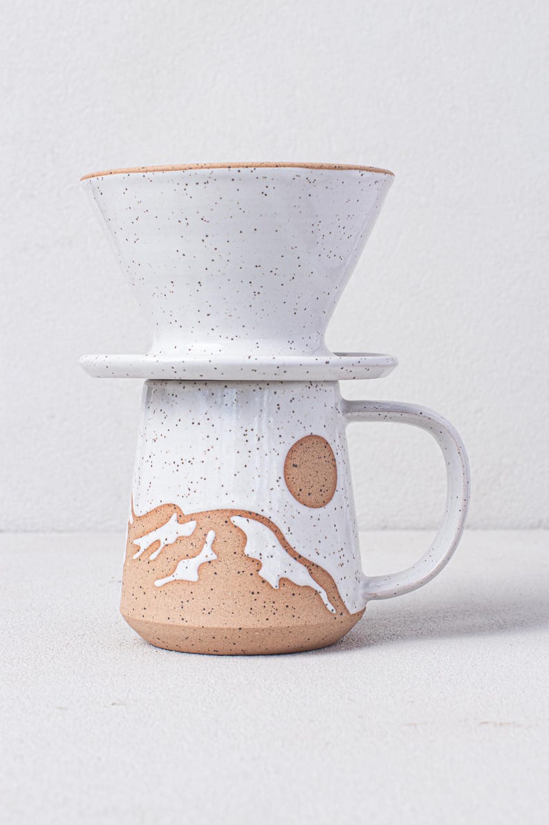 May include: White ceramic coffee pour over with a brown speckled glaze and a brown and white mountain design. The pour over sits on a matching mug with a handle.