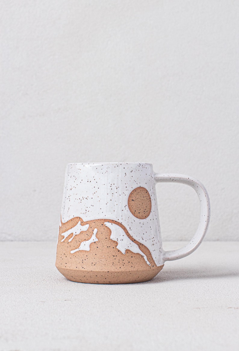May include: A white ceramic mug with a speckled glaze and a brown mountain scene design. The mug has a brown handle and a brown circle representing the sun.