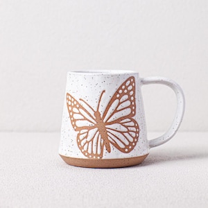 May include: White ceramic mug with a brown butterfly design. The mug has a speckled glaze and a rounded shape.