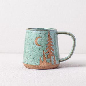 May include: A speckled teal ceramic mug with a brown base and handle. The mug features a crescent moon and tree design in brown. The mug is approximately 4 inches tall.