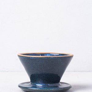 May include: A dark blue ceramic pour-over coffee dripper with a matching saucer. The dripper has a wide, shallow base and a conical shape.
