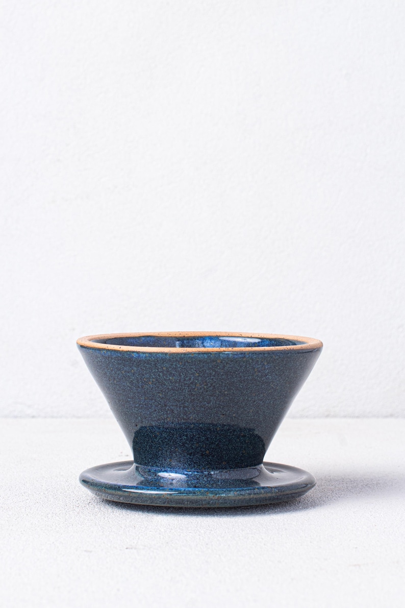 May include: A dark blue ceramic pour-over coffee dripper with a matching saucer. The dripper has a wide, shallow base and a conical shape.