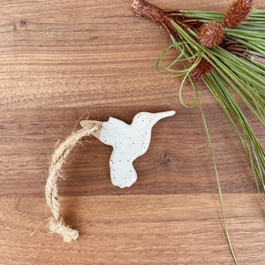 May include: A speckled white ceramic hummingbird ornament with a jute rope for hanging. The hummingbird is set against a wooden background, with a sprig of pine needles and small pine cones in the upper right corner.