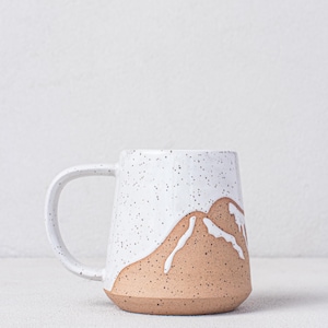 May include: A white ceramic mug with a speckled glaze and a brown mountain design. The mug has a rounded shape and a handle.