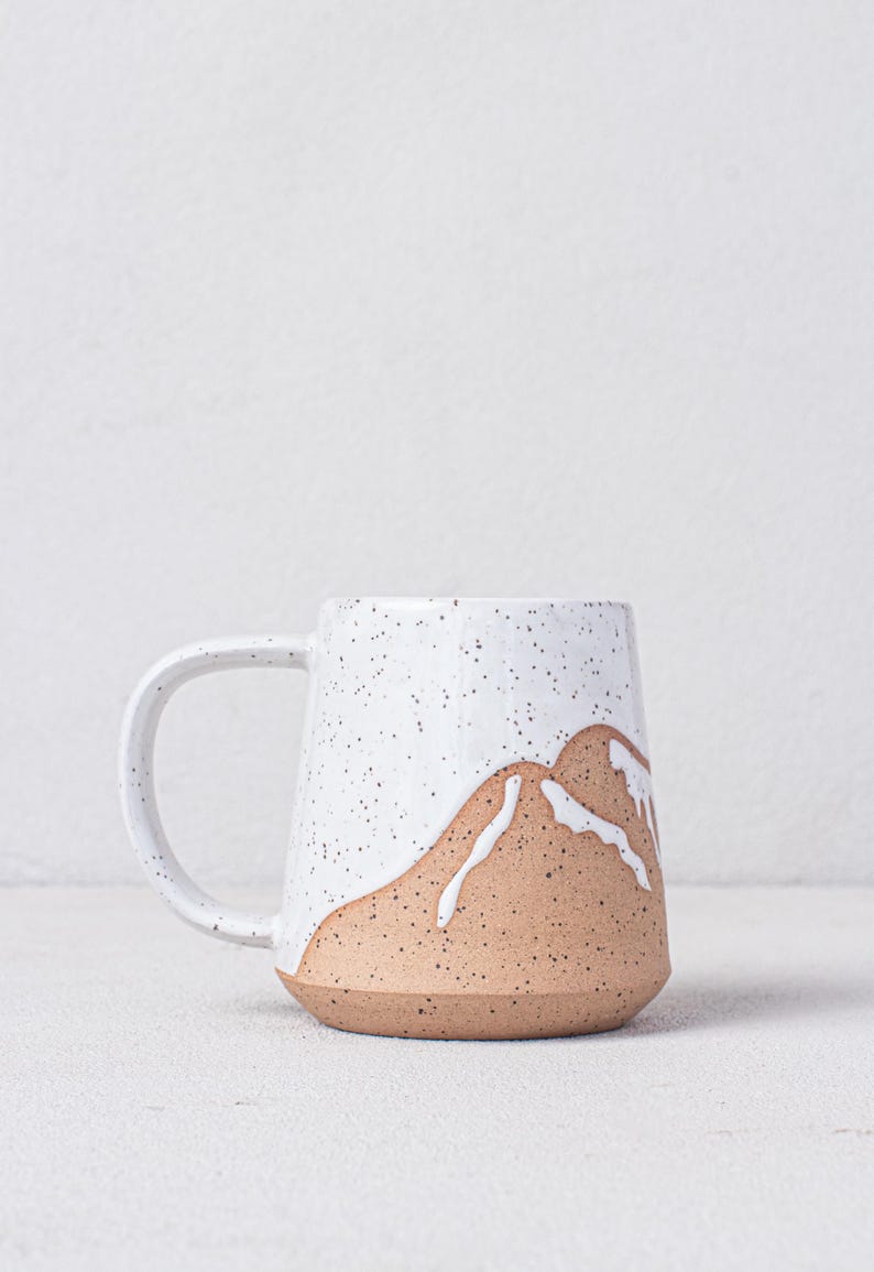 May include: A white ceramic mug with a speckled glaze and a brown mountain design. The mug has a rounded shape and a handle.