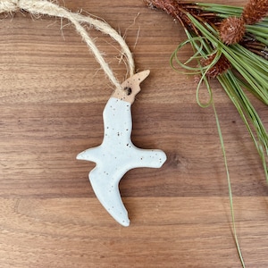 May include: A ceramic bird ornament in flight, with a speckled white body and a natural twine hanger. The bird has a tan head and is displayed on a wooden surface with pine needles and cones.