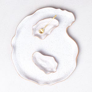 May include: A white ceramic serving tray with a speckled glaze and an irregular, organic shape. Two smaller white ceramic bowls are placed on the tray, one with a gold spoon inside.