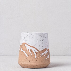 May include: A white ceramic mug with a speckled glaze and a brown mountain design. The mug is a light brown color with a white glaze on the top half. The mountain design is in white and is a simple, abstract design.