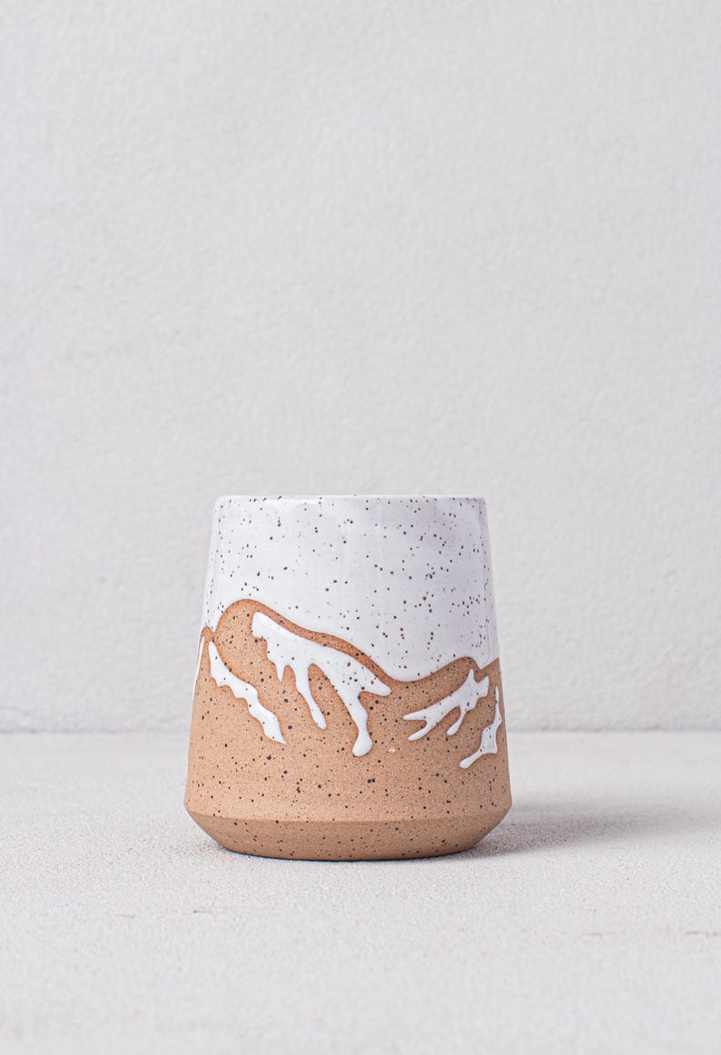 May include: A white ceramic mug with a speckled glaze and a brown mountain design. The mug is a light brown color with a white glaze on the top half. The mountain design is in white and is a simple, abstract design.