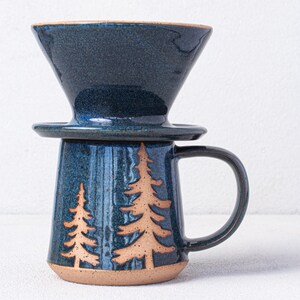 May include: A blue ceramic coffee mug with a handle and a matching blue ceramic pour-over coffee filter on top. The mug has a design of two brown trees on the front.