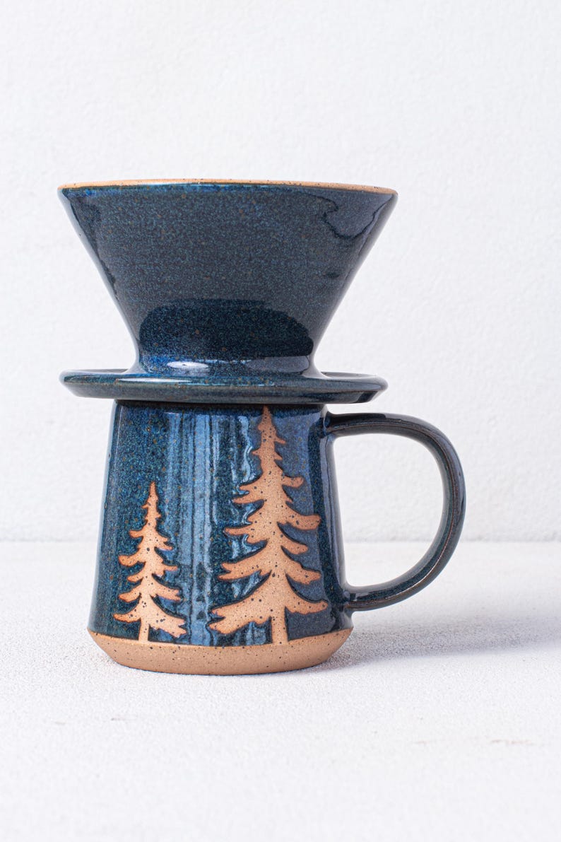 May include: A blue ceramic coffee mug with a handle and a matching blue ceramic pour-over coffee filter on top. The mug has a design of two brown trees on the front.