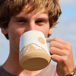May include: A white ceramic mug with a brown speckled glaze. The mug features a white mountain range design with a brown sun and waves.